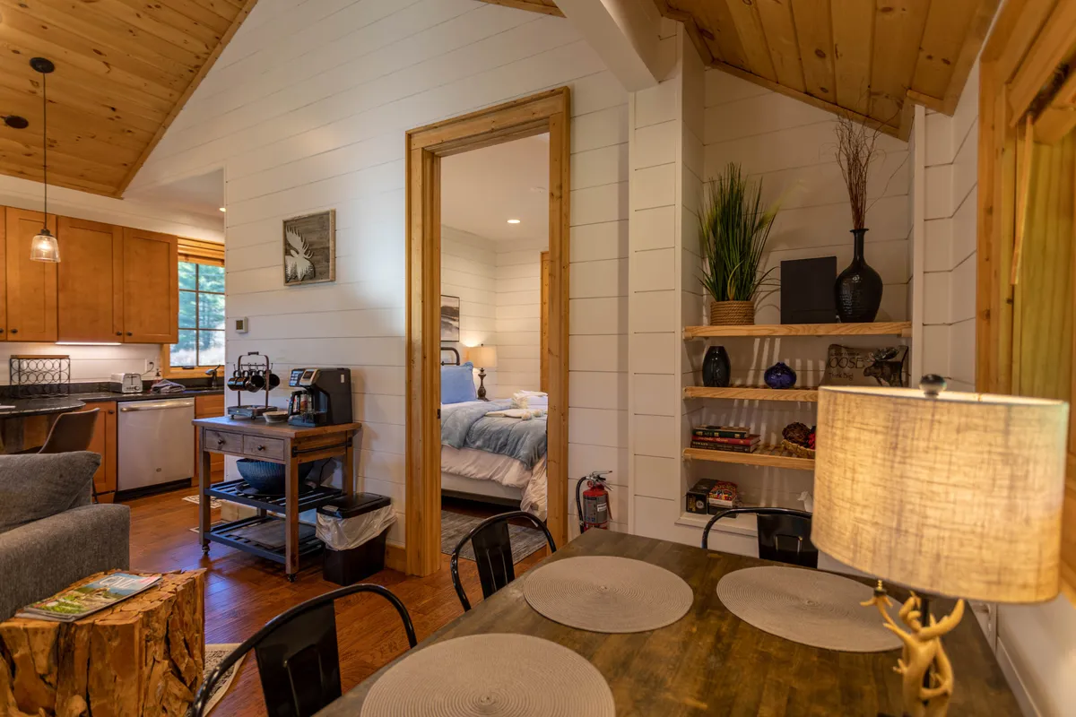 A bright, inviting open-plan space featuring a dining table set beside a window, a well-equipped kitchen, a glimpse into a cozy bedroom, and a bookshelf. The large windows bring in natural light and hint at views of the outdoors, creating a sense of connection with the surrounding landscape.