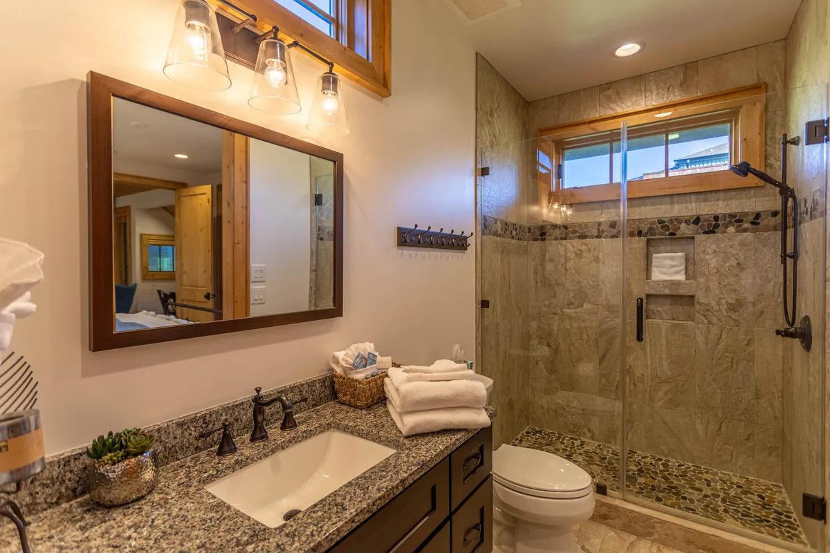 A modern bathroom with a striking granite countertop, large mirror, glass shower enclosure, and wooden accents. Sunlight streams in, enhancing the clean, luxury feel of the space.