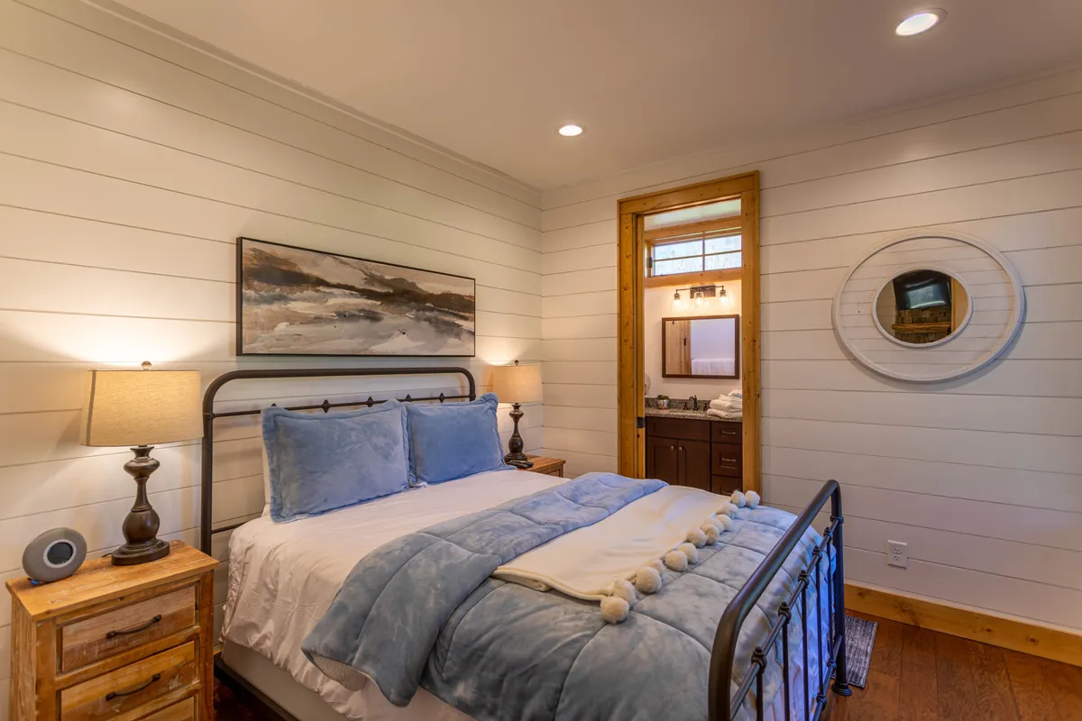 A comfortable bedroom with blue bedding, framed artwork, a nightstand with a lamp, and a decorative mirror. The soft lighting and cohesive decor create a peaceful, cozy ambiance.