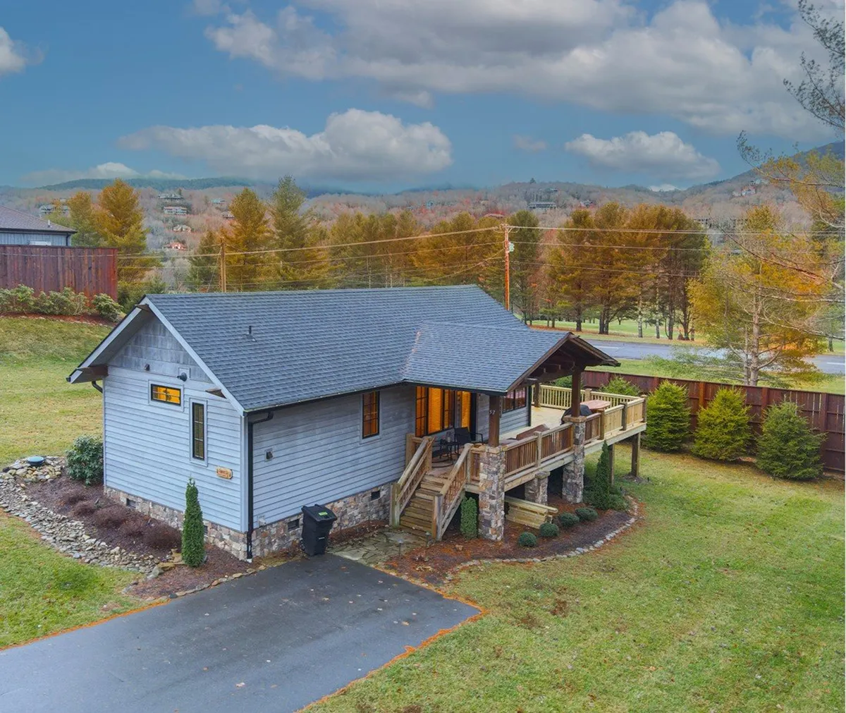 The image showcases the charming cabin exterior with a spacious wooden wrap-around porch, set against a breathtaking backdrop of rolling North Carolina mountains. The setting sun casts a warm glow over the scene, inviting guests to relax outdoors and soak in the tranquil surroundings.