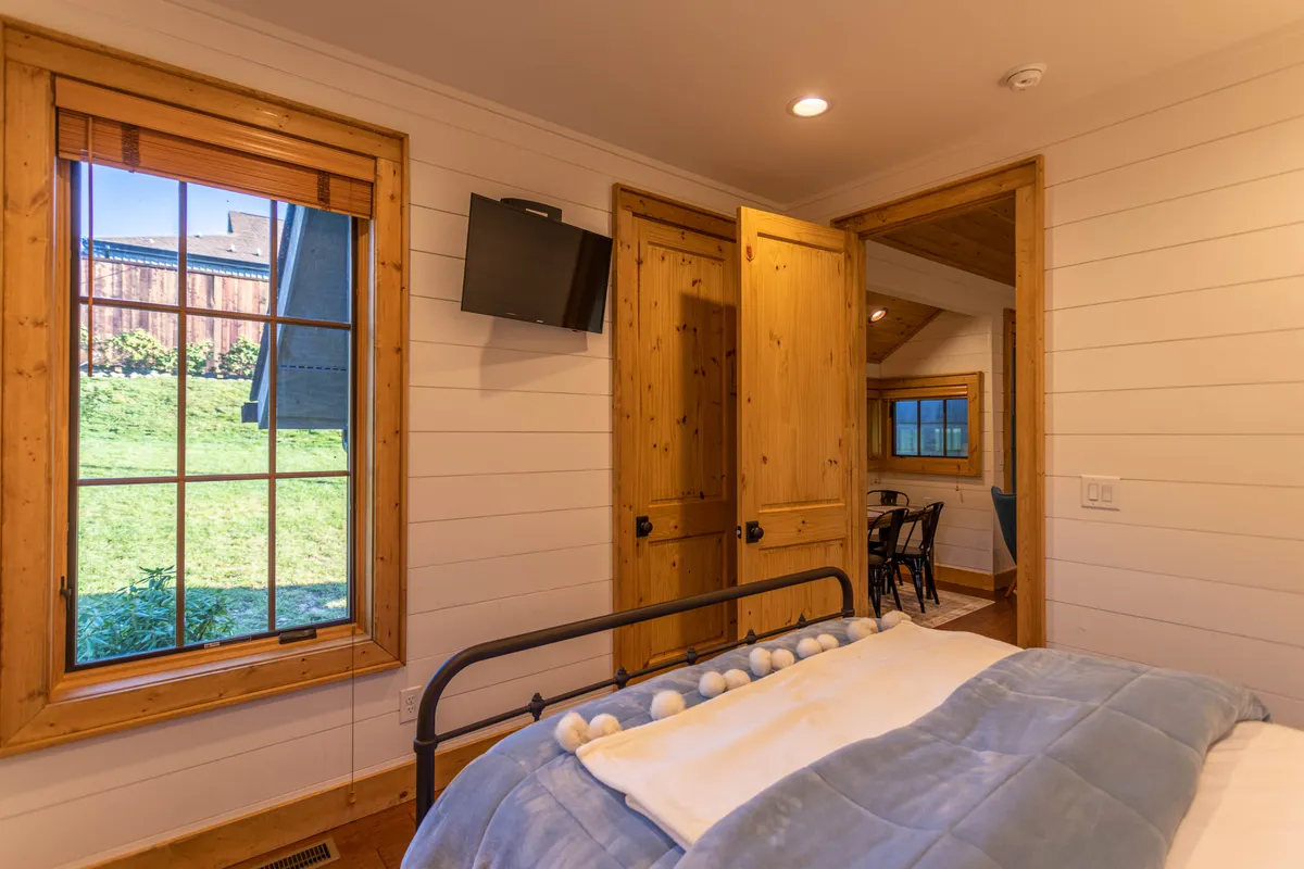 A cozy bedroom scene with a bed dressed in soft bedding, flanked by wooden doors and a large window that opens up to the outside world. The room is filled with natural light, evoking comfort and a gentle start to the day.