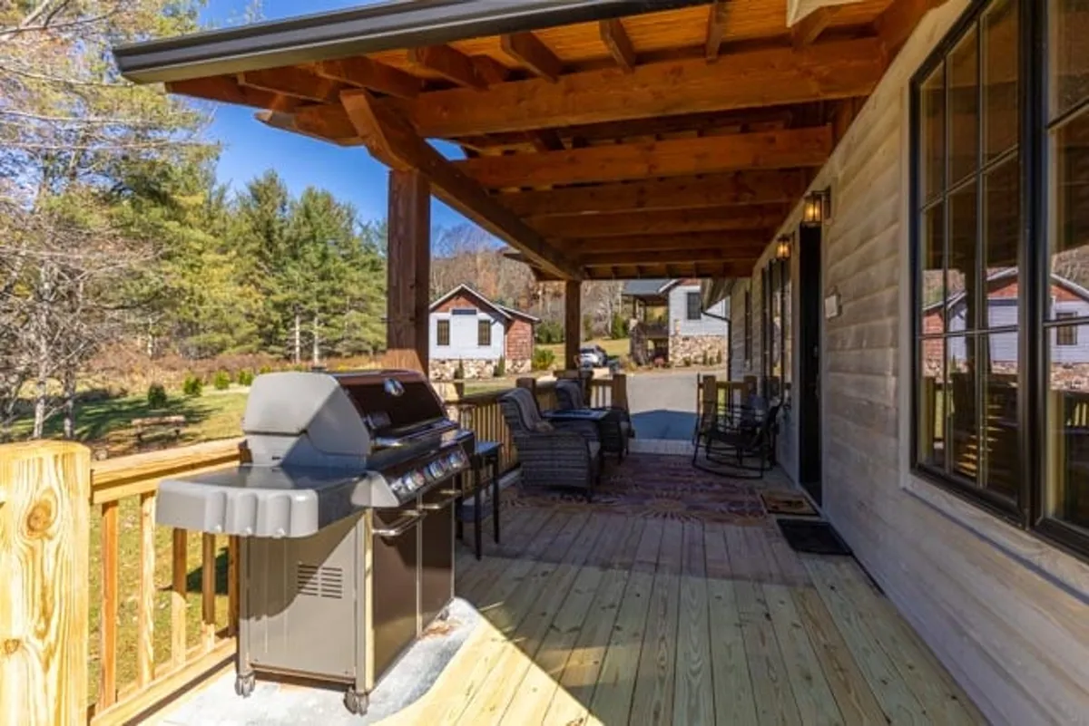 This exterior image showcases a wooden deck surrounded by trees, featuring a grill and comfortable outdoor seating. The setting is inviting, with natural sunlight filtering through the leaves, making it an ideal space for guests to unwind, share meals, and enjoy the mountain air.