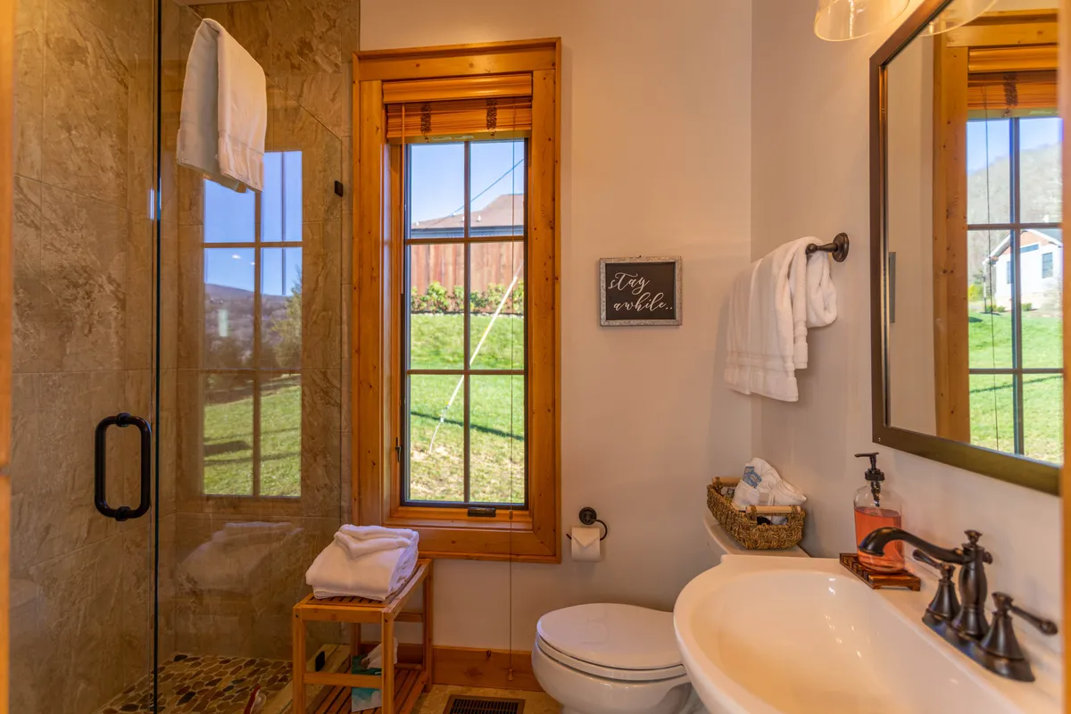 A serene shower area featuring a large window that frames a tranquil view of the outdoors, accompanied by a decorative wall sign and a well-appointed sink area. Natural light pours in, suggesting a seamless connection to the surrounding environment and an invitation to relax in privacy.