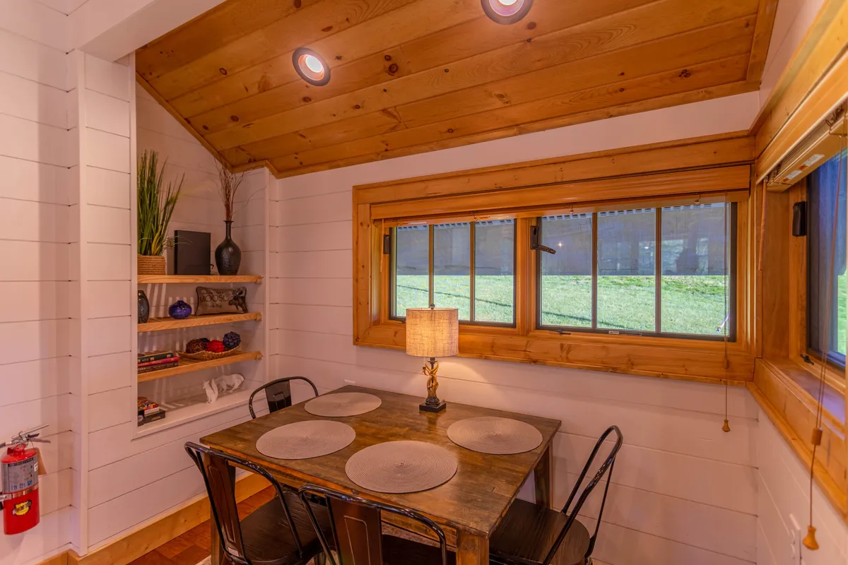 A cozy wooden table sits near a large window, inviting couples to share a meal or a cup of coffee while enjoying the view. Warm lighting from a table lamp and tasteful decorative shelving create an inviting, intimate ambiance. The window hints at the scenic outdoors, subtly connecting the interior with the vibrant landscape beyond.