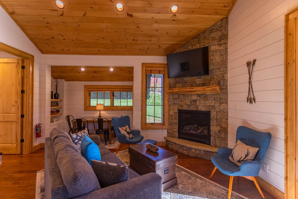 A warm and welcoming living room centered around a striking stone gas log fireplace. Comfortable seating faces both the fireplace and a large window, which bathes the room in natural light and offers glimpses of the mountain landscape. The space feels perfect for relaxing, socializing, or enjoying a movie night.
