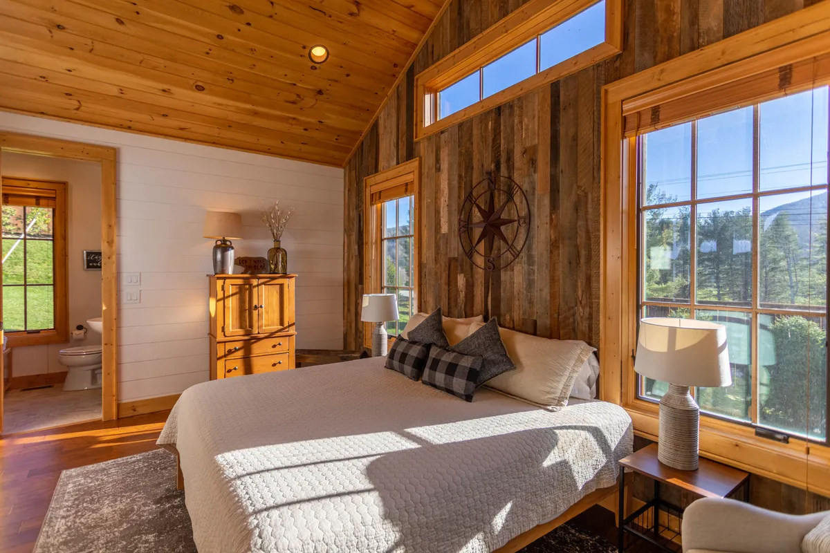 A serene bedroom is bathed in morning light through large windows that look out onto the natural surroundings. Decorative pillows and a classic wooden dresser add luxury, while the windows offer glimpses of the tranquil, leaf-filled landscape.