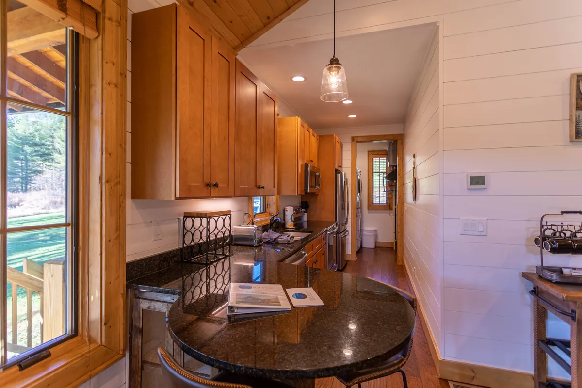 A view of the open kitchen featuring granite countertops, wooden cabinets, large sunny windows, and bar stools for casual dining or conversation. The setting is staged for cooking and socializing, with ample space for multiple guests to participate in meal prep or sit and chat.