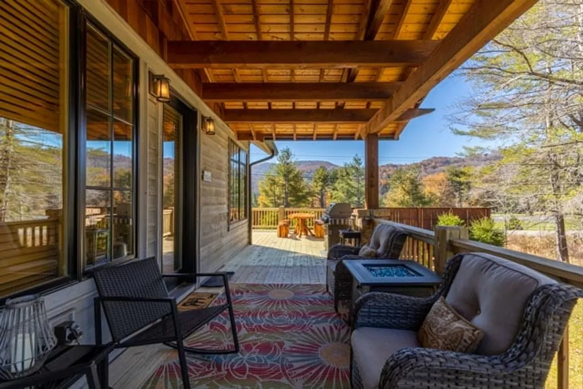 A cozy, comfortable seating area is arranged on a wraparound porch space, complete with a decorative rug and panoramic mountain views beyond the railing. The scene captures the warmth and inviting atmosphere ideal for morning coffee or evening gatherings.