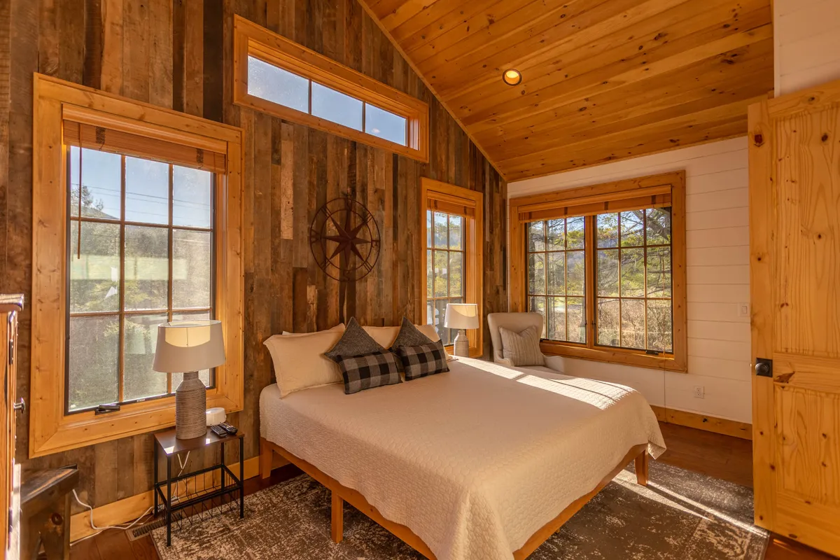 A serene bedroom featuring a plush bed adorned with decorative pillows, warm wooden wall paneling, and large windows that flood the space with natural light. The inviting atmosphere is enhanced by the view outside and a sense of cozy relaxation.