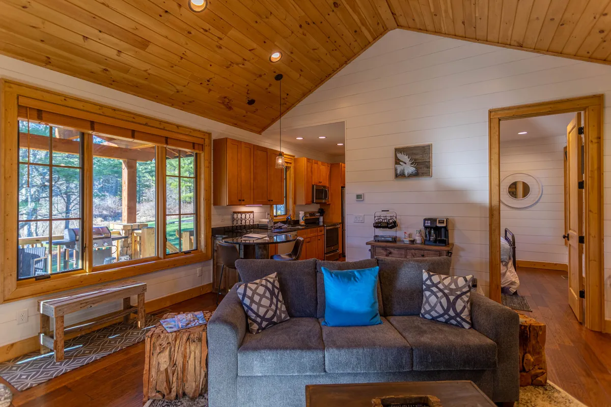 An open-plan living area highlighting a comfortable sofa with a view through a large window, blending seamlessly into the kitchen space. The room is bathed in natural light, making it ideal for both relaxation and socializing, whether you're watching the sunrise or preparing a meal.